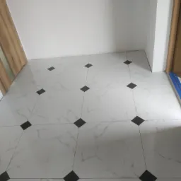 Wykończona podłoga z białych płytek imitujących marmur z czarnymi wstawkami w kształcie rombów, widoczny fragment drewnianej ościeżnicy drzwi i niebieska poziomica.