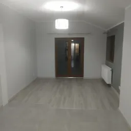 Pomalowane na szaro wnętrze pokoju z drewnianą podłogą i grzejnikiem, widok na przeszklone drzwi balkonowe.