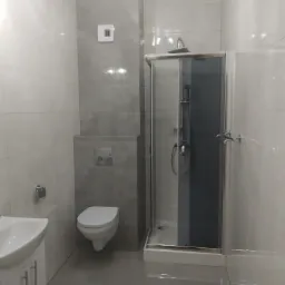 Nowoczesna łazienka z szarymi i białymi płytkami, kabiną prysznicową z grafitowym szkłem, podwieszaną toaletą i białą umywalką z szafką.