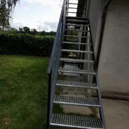 Zewnętrzne metalowe schody z kratowymi stopniami, prowadzące do dachu budynku, na tle zielonego trawnika i krzewów.