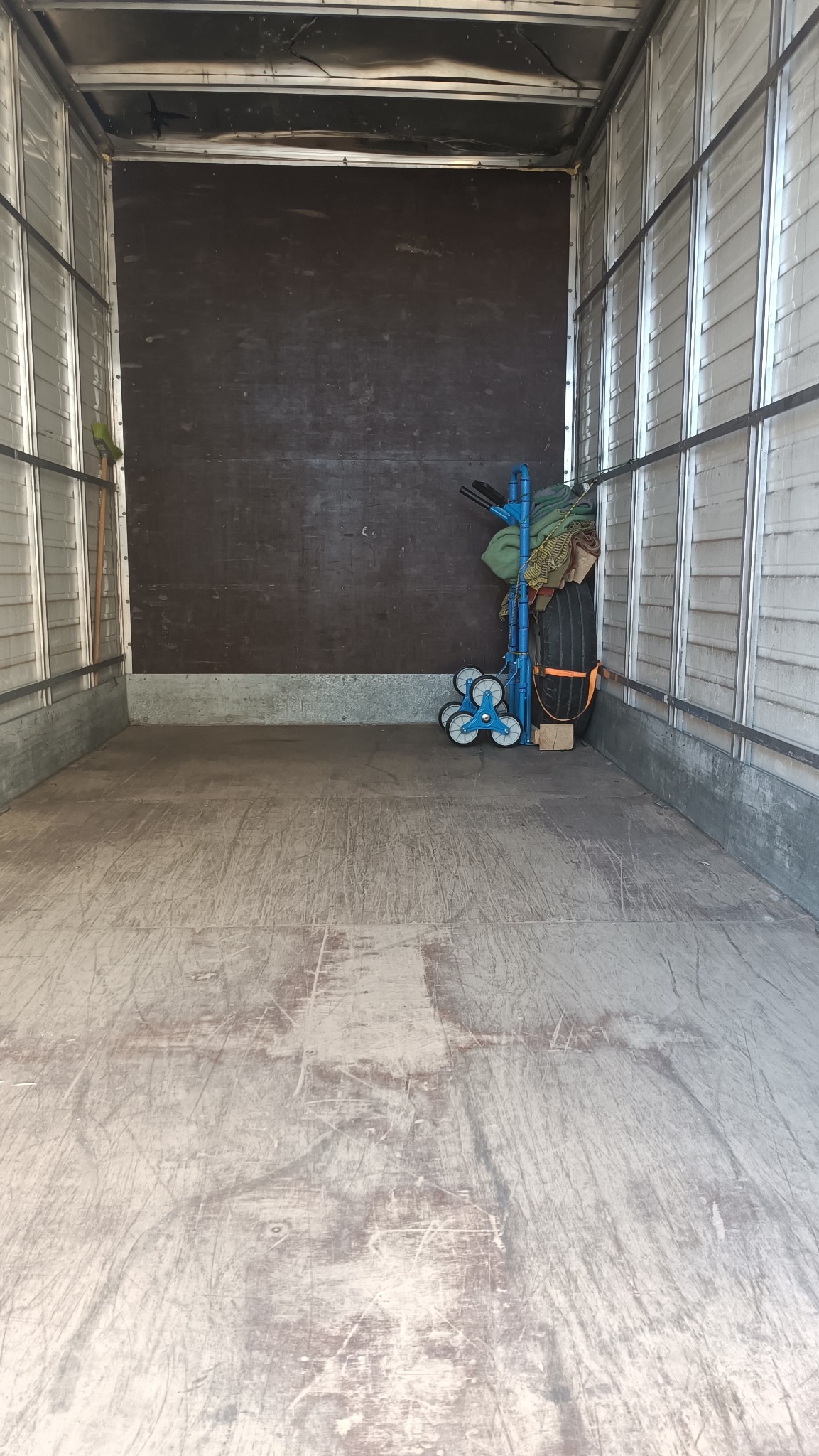Wnętrze paki busa z widoczną ścianą z płyty, podłogą zarysowaną śladami użytkowania oraz wózkiem transportowym z oponą i zabezpieczeniami w rogu.