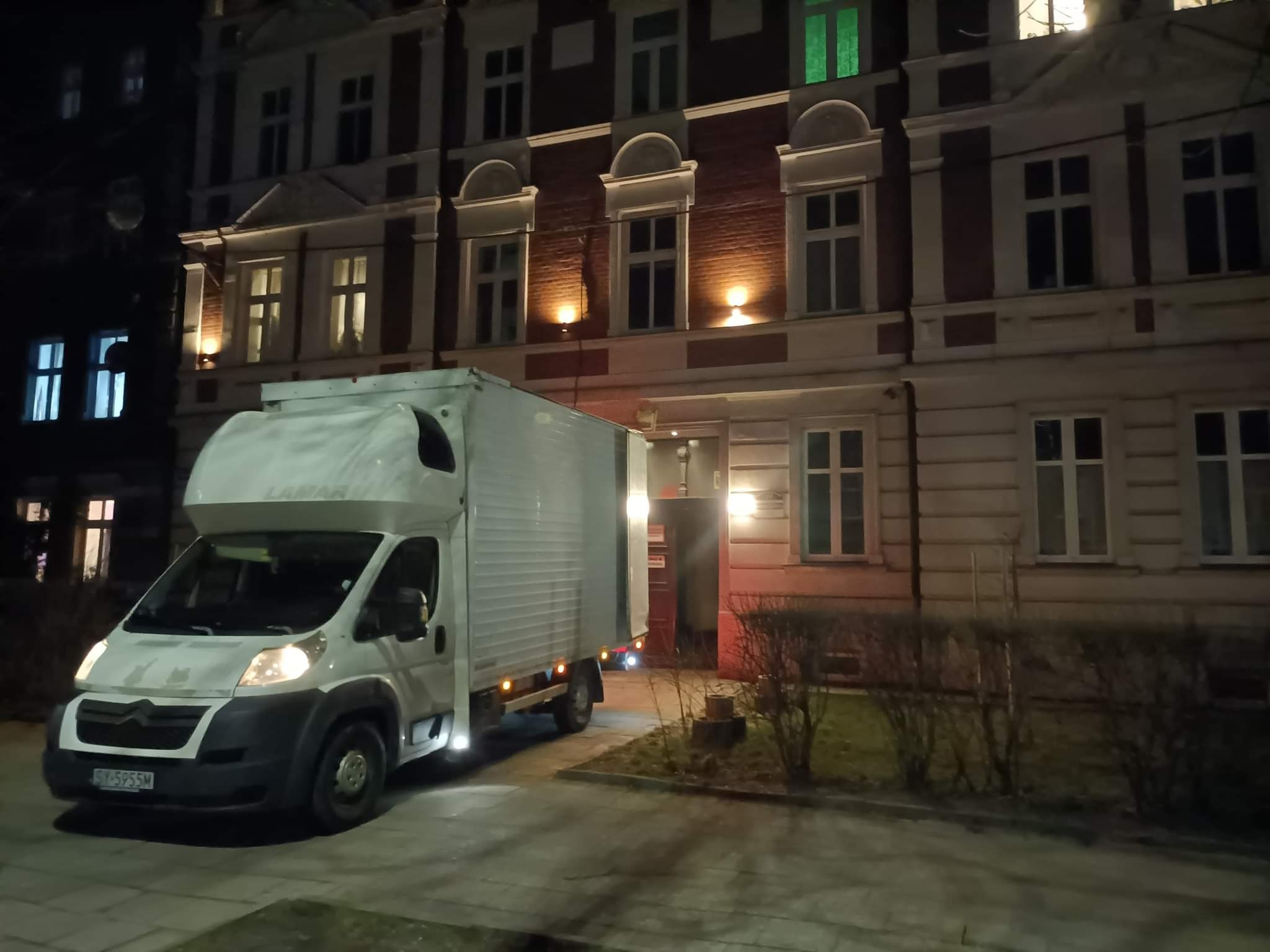 Biały samochód dostawczy zaparkowany przed kamienicą w nocy, widoczne światła w oknach i tablica rejestracyjna pojazdu.