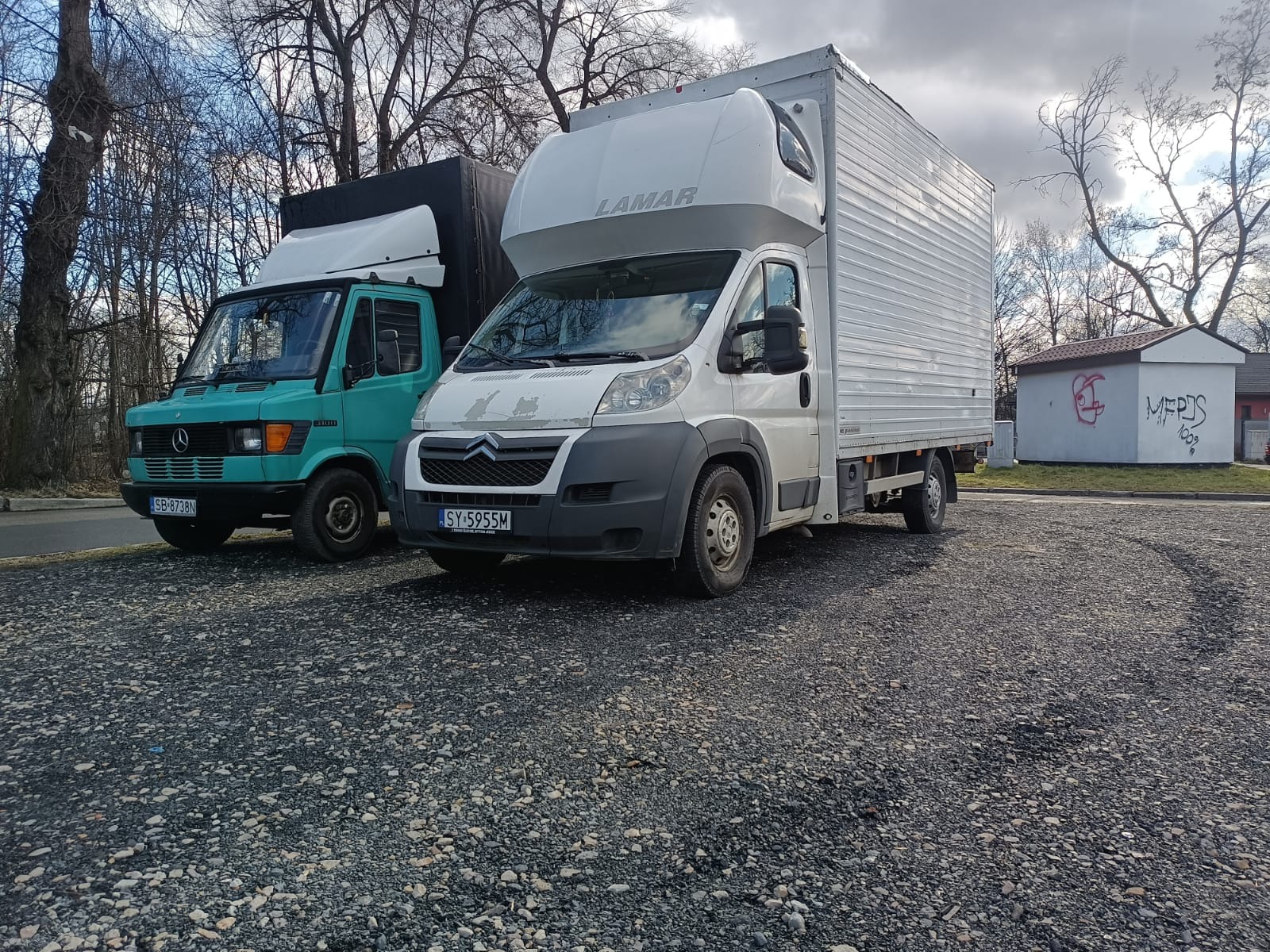 Dwa samochody dostawcze zaparkowane na żwirowym parkingu, w tle budynek z graffiti i nagie drzewa.