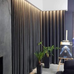 Edith Styl - Długie, grafitowe zasłony z ukrytym oświetleniem LED u góry, dwie rośliny doniczkowe w czarnych, kwadratowych donicach, obraz na sztaludze w tle, szara ściana z efektem betonu i fragment czarnej sofy.