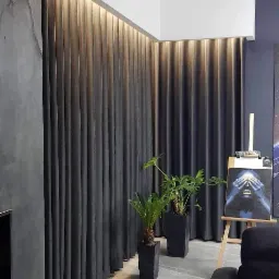 Długie, grafitowe zasłony z ukrytym oświetleniem LED u góry, dwie rośliny doniczkowe w czarnych, kwadratowych donicach, obraz na sztaludze w tle, szara ściana z efektem betonu i fragment czarnej sofy.