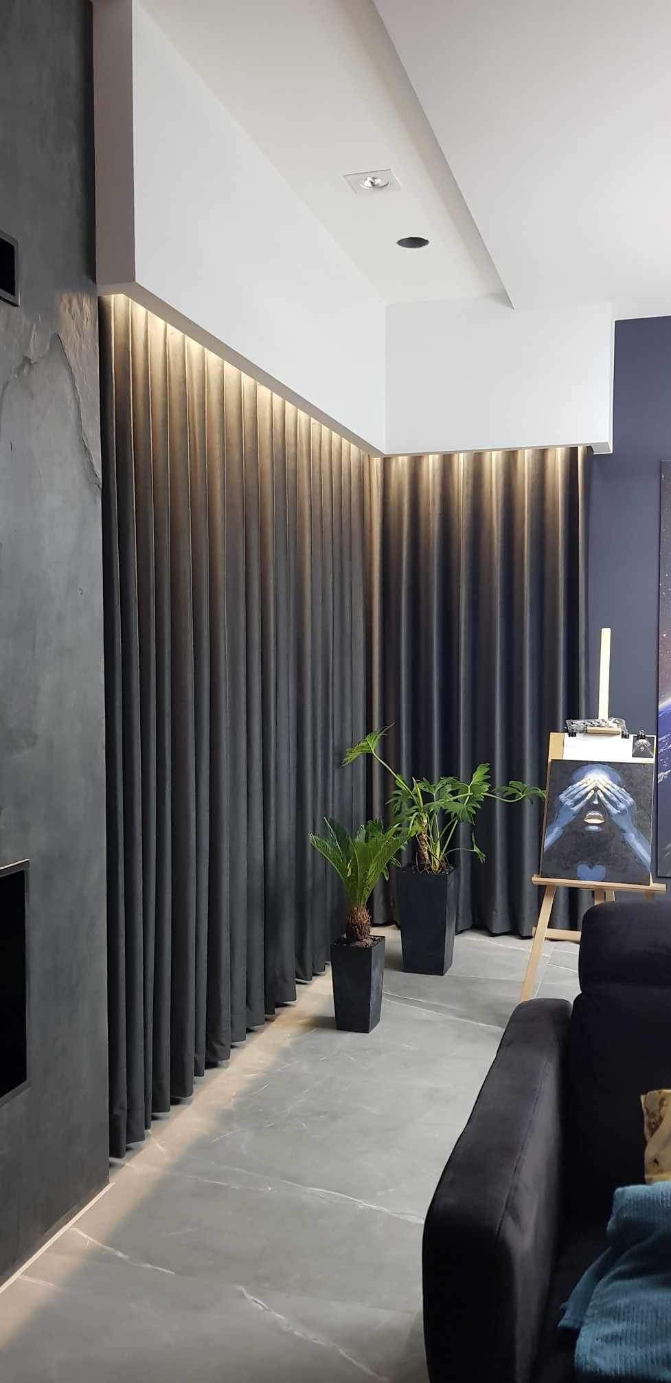 Długie, grafitowe zasłony z ukrytym oświetleniem LED u góry, dwie rośliny doniczkowe w czarnych, kwadratowych donicach, obraz na sztaludze w tle, szara ściana z efektem betonu i fragment czarnej sofy.