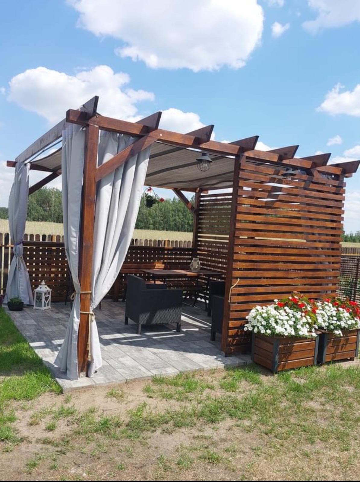 Drewniana pergola z szarymi zasłonami, oświetleniem i skrzyniami kwiatów na zewnątrz, na tle pola i błękitnego nieba.