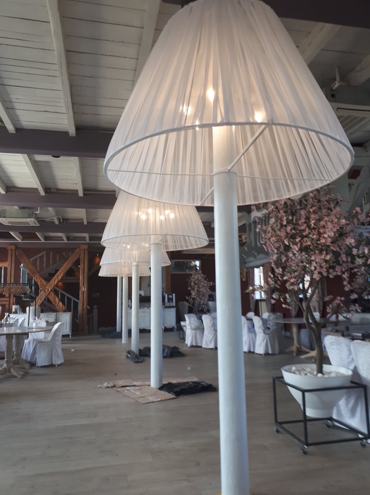 Sala bankietowa z rzędem wysokich lamp podłogowych z abażurami z białej, prześwitującej tkaniny i dekoracjami z kwitnących gałązek, przygotowywana do uroczystości.