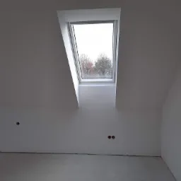 Poddasze z oknem dachowym po szpachlowaniu, widoczne otwory na gniazdka elektryczne i surowa podłoga.