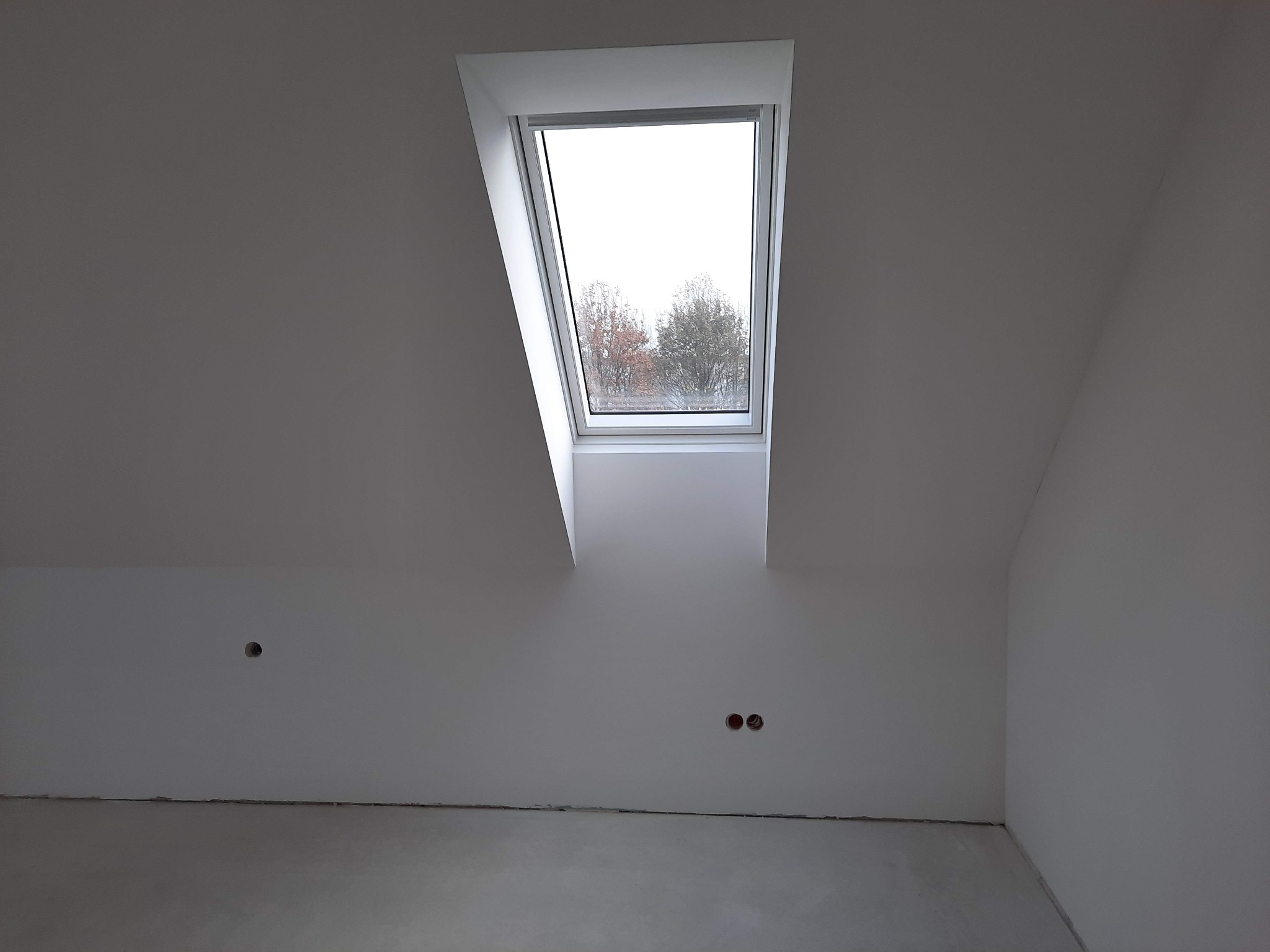 Poddasze z oknem dachowym po szpachlowaniu, widoczne otwory na gniazdka elektryczne i surowa podłoga.