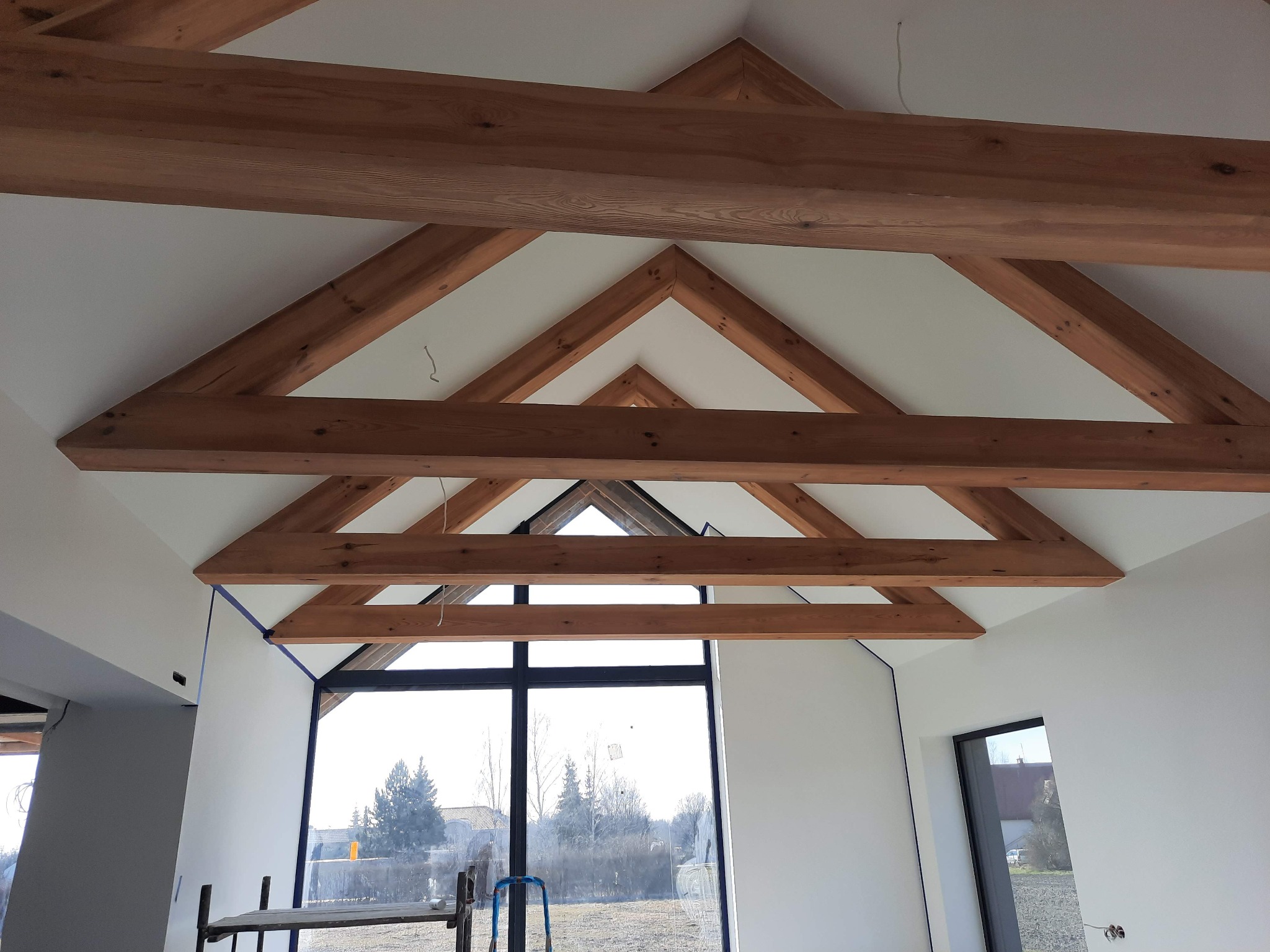 Wykończone poddasze z widoczną drewnianą konstrukcją więźby dachowej, białe ściany pomalowane na gładko, duże okno z widokiem na zewnątrz, zabezpieczone taśmą malarską krawędzie.