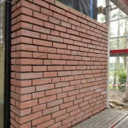 Ściana z czerwonej cegły klinkierowej w trakcie murowania, widoczny narożnik i fragment rusztowania.