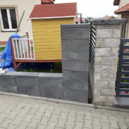 Wznoszenie muru oporowego z szarych bloczków betonowych, obok drewniany domek dla dzieci z czerwonym dachem i fragment murowanego słupka ogrodzeniowego z jasnego kamienia.