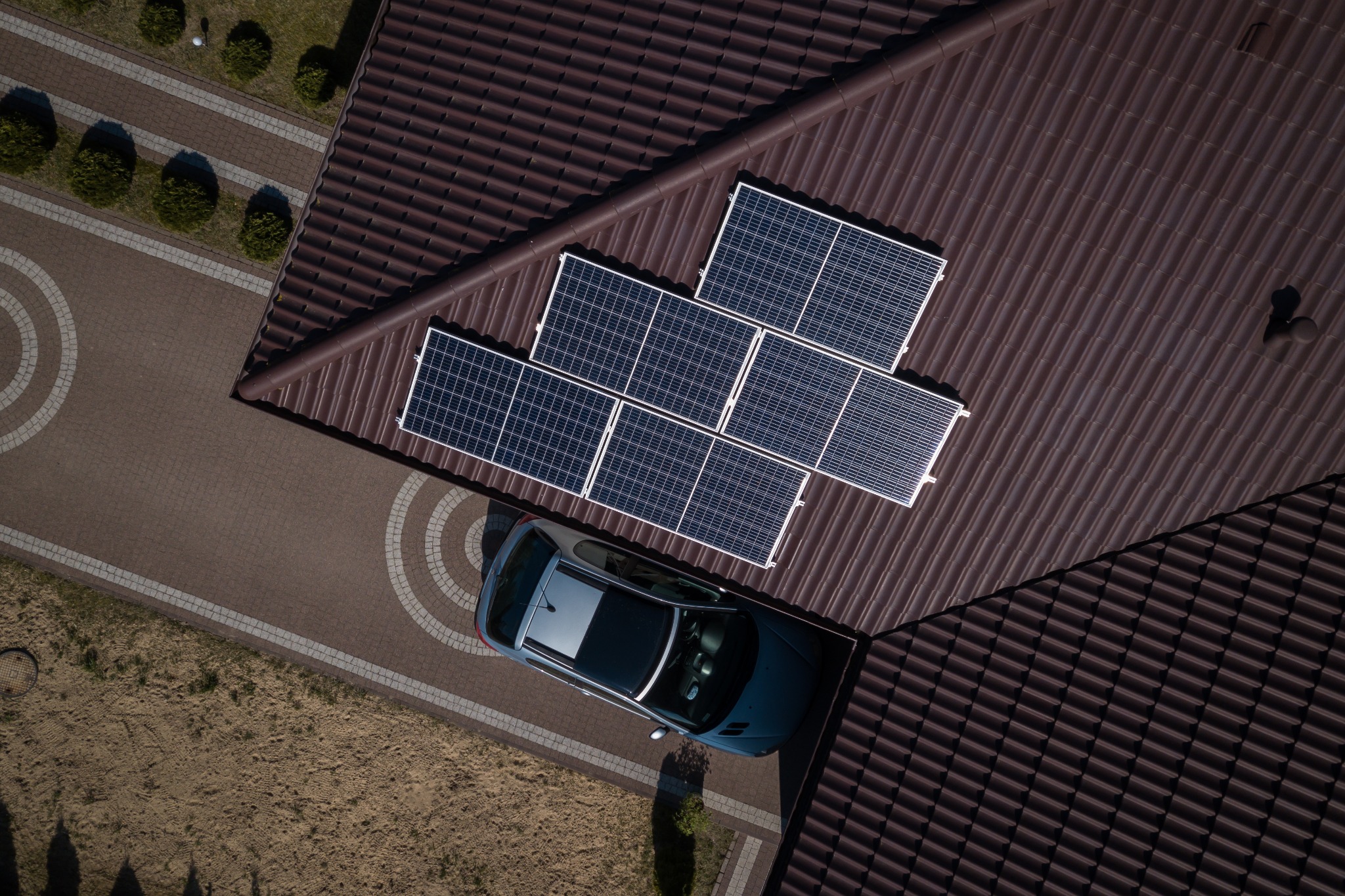 Widok z góry na brązowy dach z zamontowanymi panelami słonecznymi, obok zaparkowany szary samochód na brukowanym podjeździe.