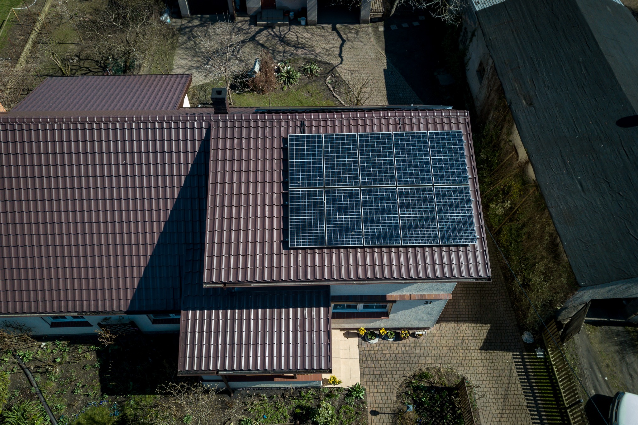 Widok z góry na brązowy dach domu z zainstalowanymi panelami słonecznymi w układzie 2x4, obok widoczny ogród i chodnik z kostki.