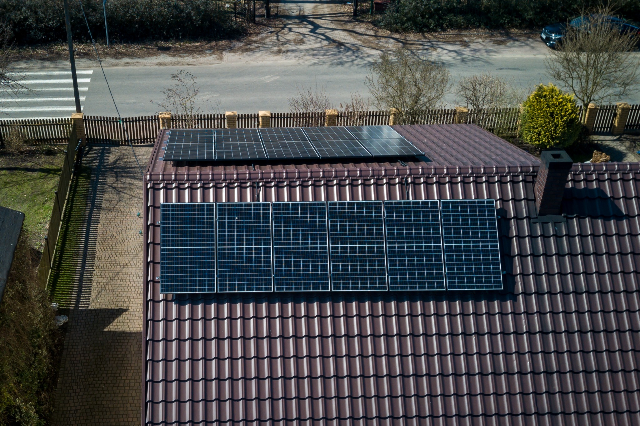 Widok z góry na brązowy dach z dachówki z dwoma rzędami paneli fotowoltaicznych; jeden rząd z pięcioma panelami, drugi z trzema, z kominem w tle i widoczną ulicą z przejściem dla pieszych.