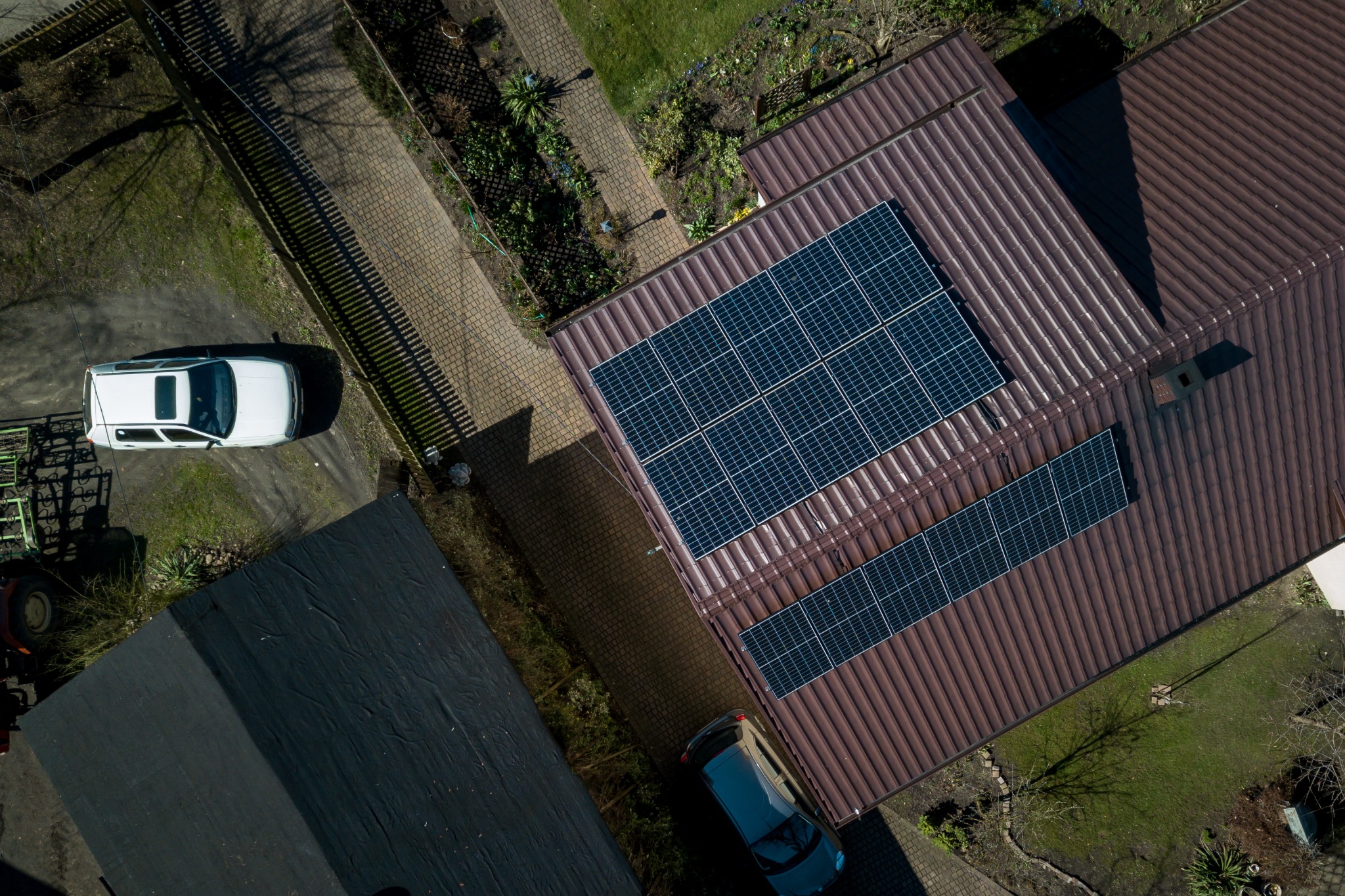 Widok z góry na brązowy dach z zainstalowanymi panelami słonecznymi, obok zaparkowany biały samochód i zadbany ogród.