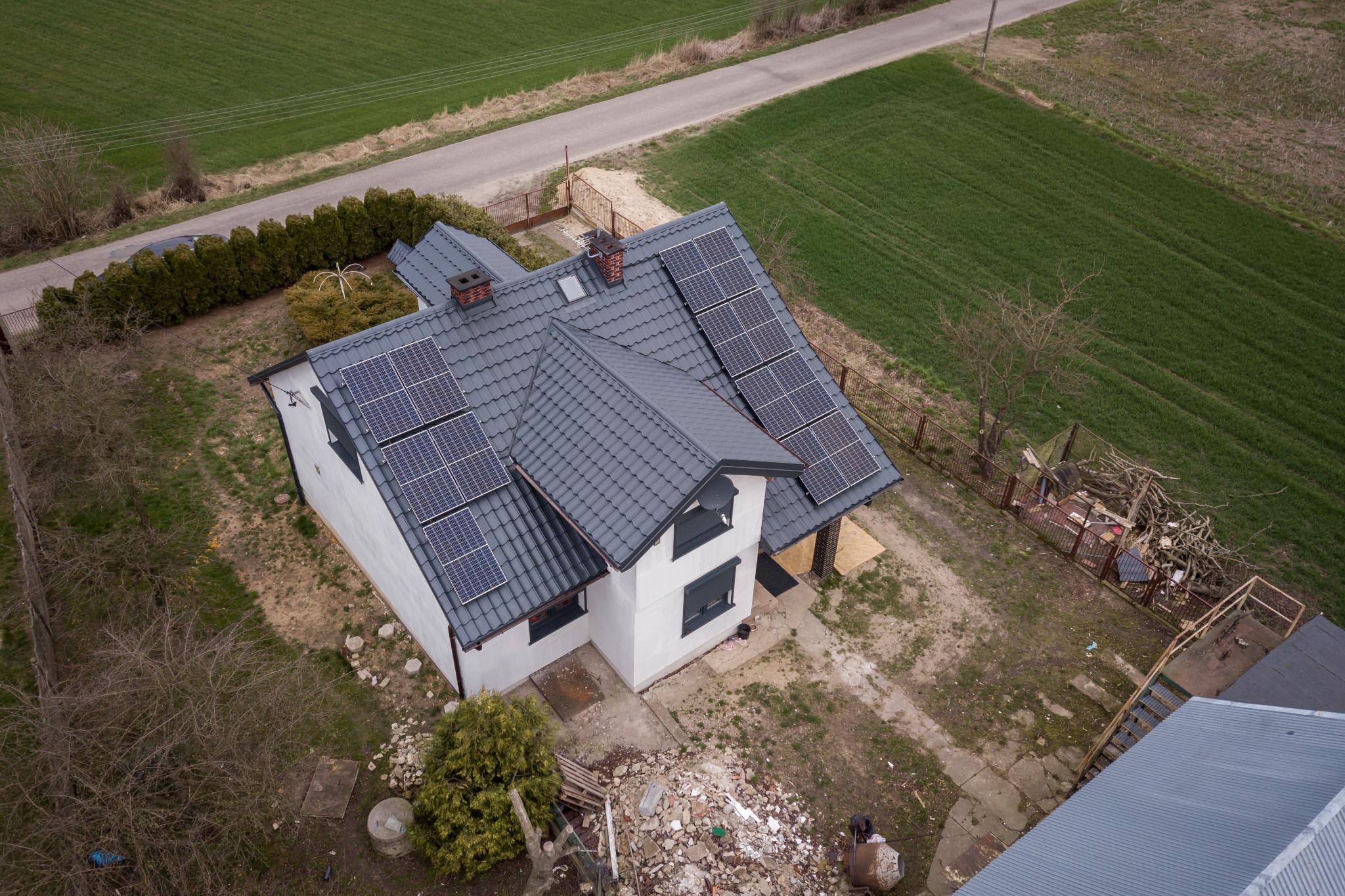 Widok z góry na dom jednorodzinny z szarym dachem, na którym zamontowano panele słoneczne; w tle zielone pola i droga.