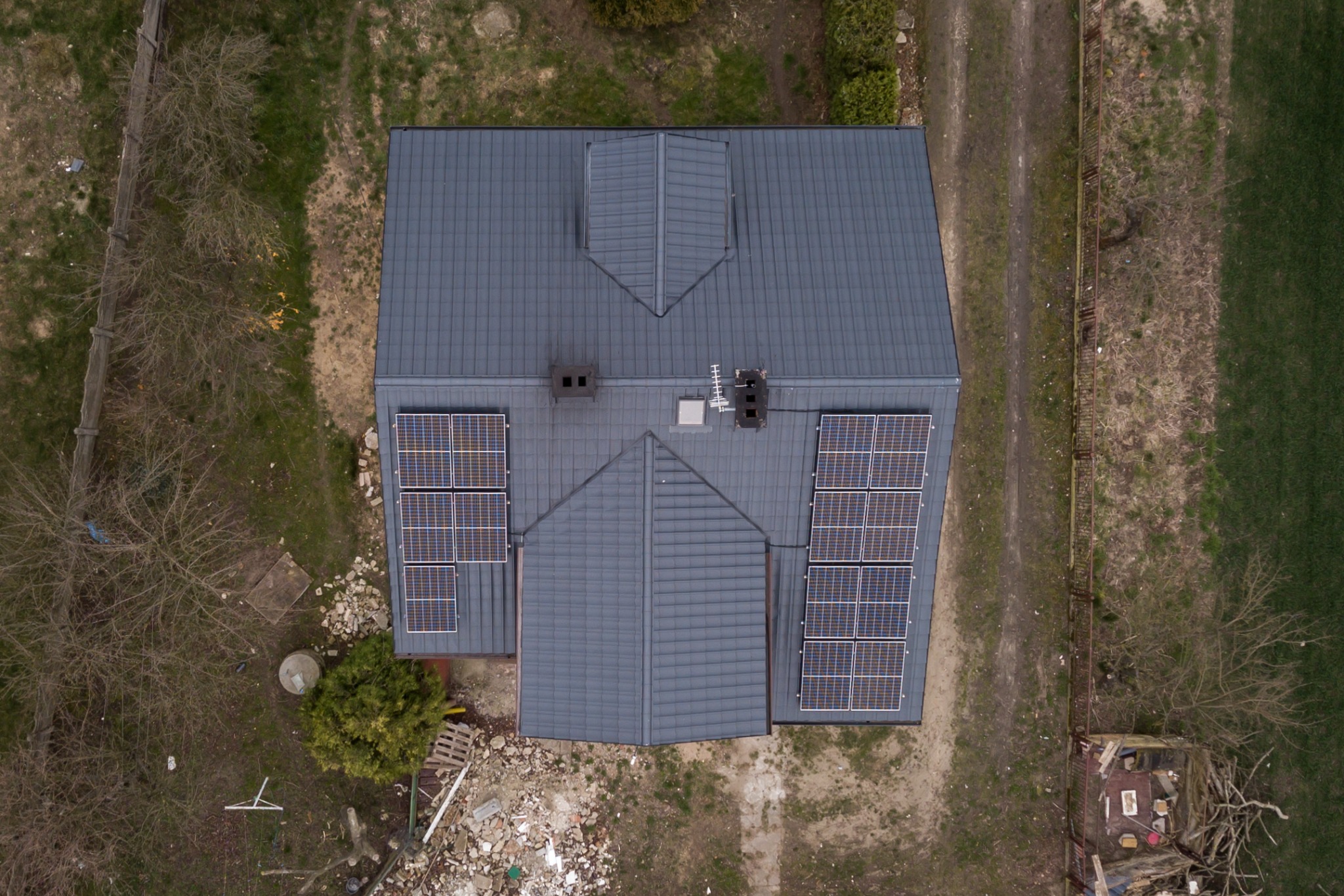 Widok z drona na szary dach domu w Rzgowie z panelami słonecznymi, otoczonego drzewami i niezagospodarowanym terenem.