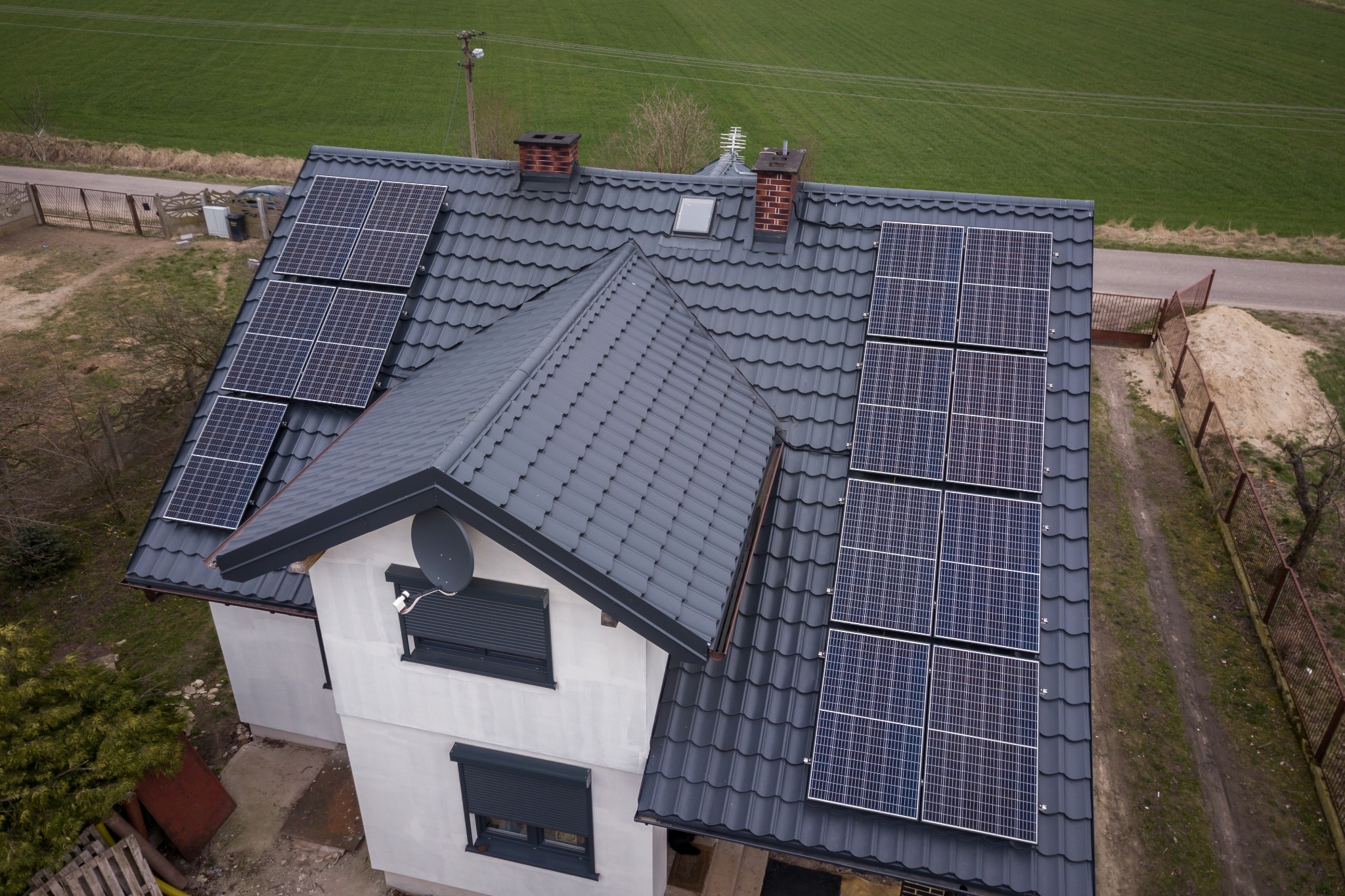 Widok z góry na dom z szarą dachówką i panelami słonecznymi, z anteną satelitarną na bocznej ścianie i polem w tle.