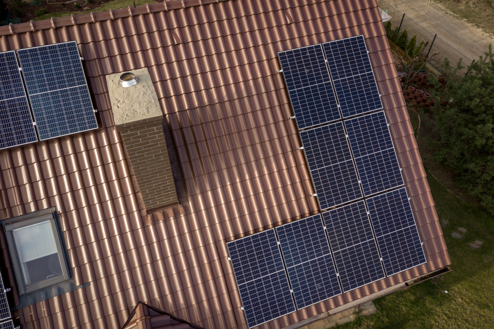Widok z góry na brązowy dach z dachówki ceramicznej z zainstalowanymi panelami fotowoltaicznymi i kominem z cegły klinkierowej.