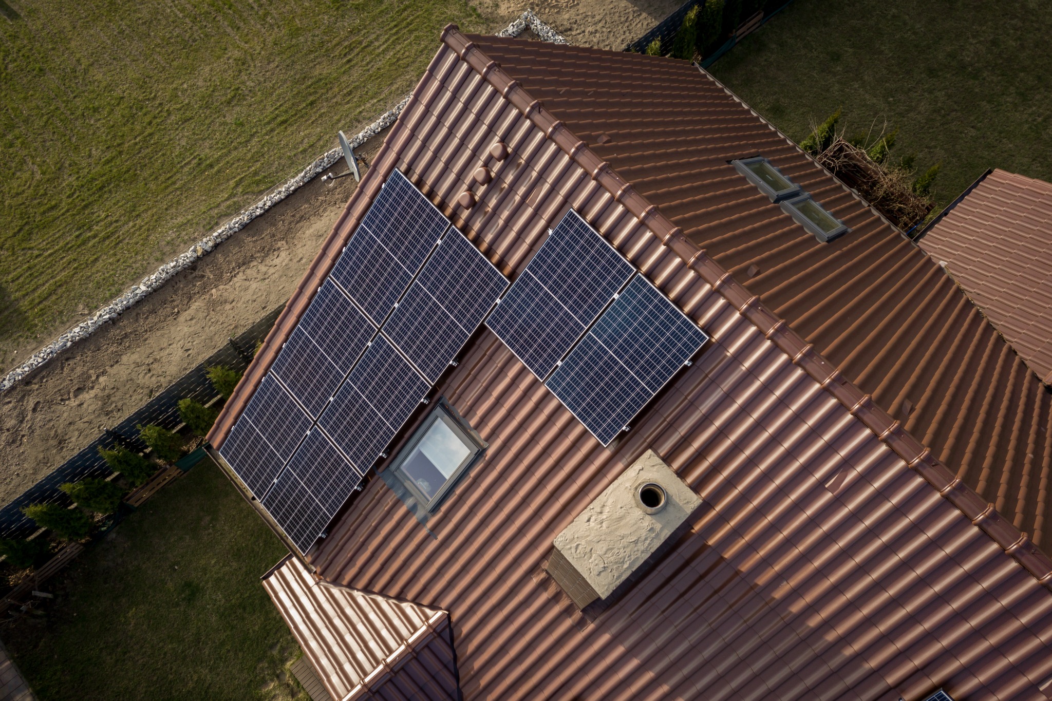 Widok z góry na brązowy dach z panelami słonecznymi, kominem i dwoma oknami dachowymi, otoczony zielonym trawnikiem i kamiennym obrzeżem.