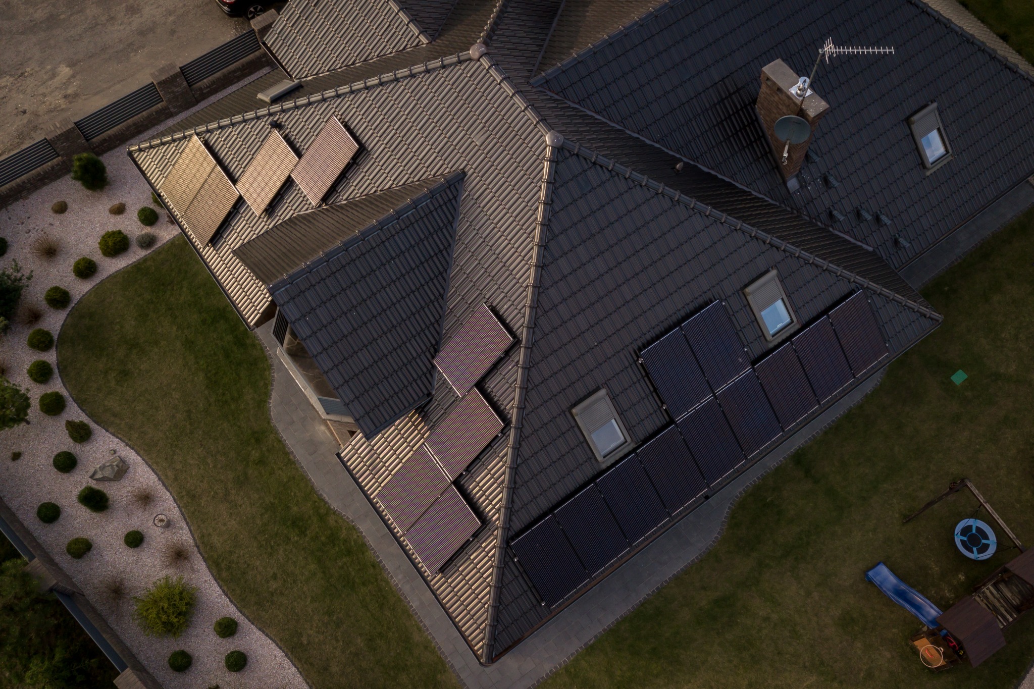 Widok z góry na dach domu z ciemnej dachówki z zamontowanymi panelami słonecznymi; część paneli odbija zachodzące słońce, na dachu widoczne okna dachowe, komin i antena.