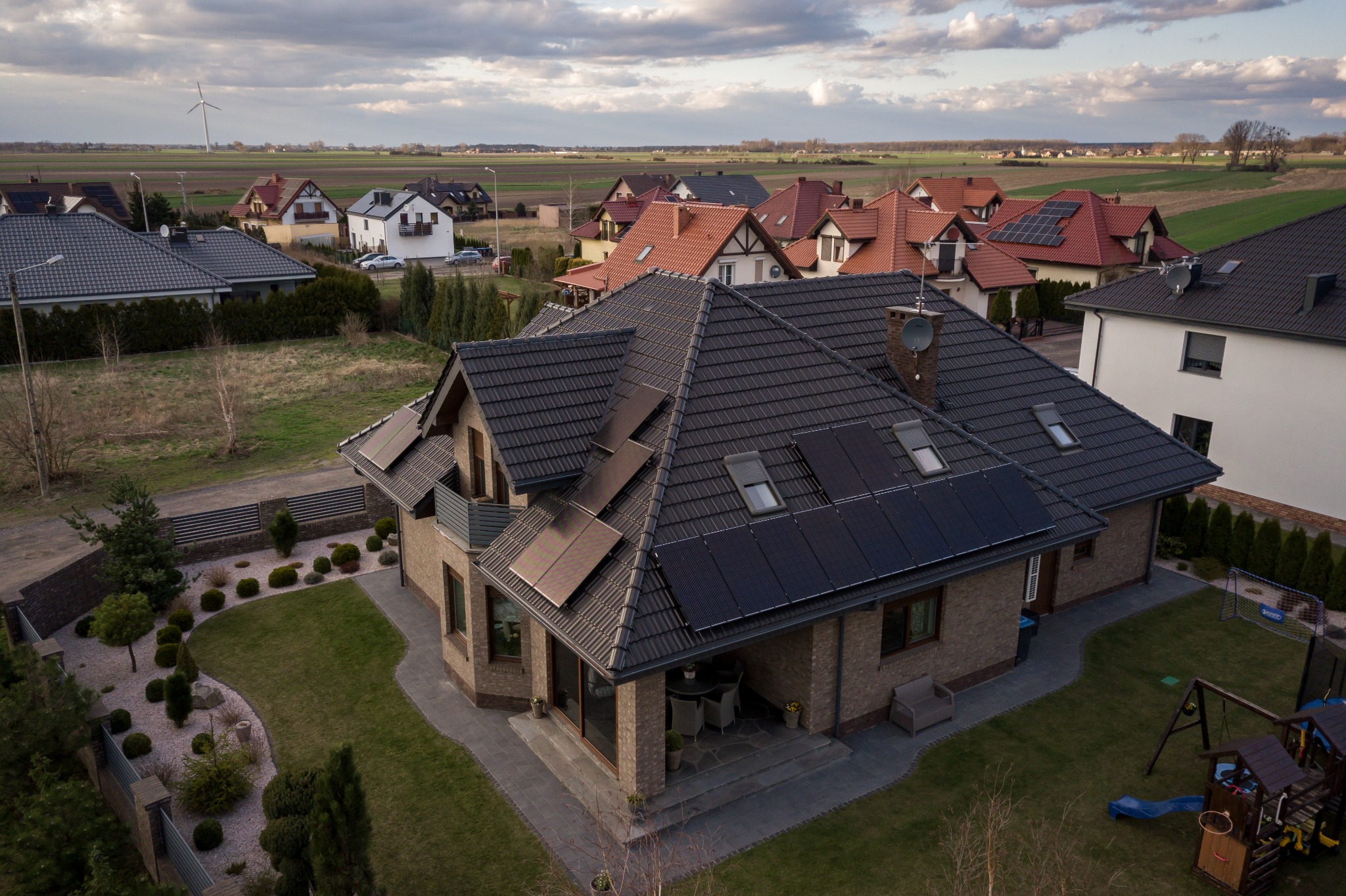 Widok z góry na dom jednorodzinny z panelami słonecznymi na dachu, w tle inne domy, pola i turbina wiatrowa.