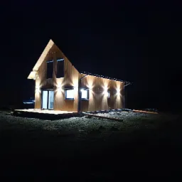 Nowoczesny dom z drewnianą elewacją oświetlony zewnętrznymi lampami w nocy, widoczne rury odprowadzające wodę.