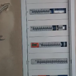 Rozdzielnica elektryczna z widocznymi bezpiecznikami i przewodami, zamontowana na ścianie z surowym wykończeniem. Widoczny licznik Simlic LS-3F. Część bezpieczników jest opisana marką Eaton.