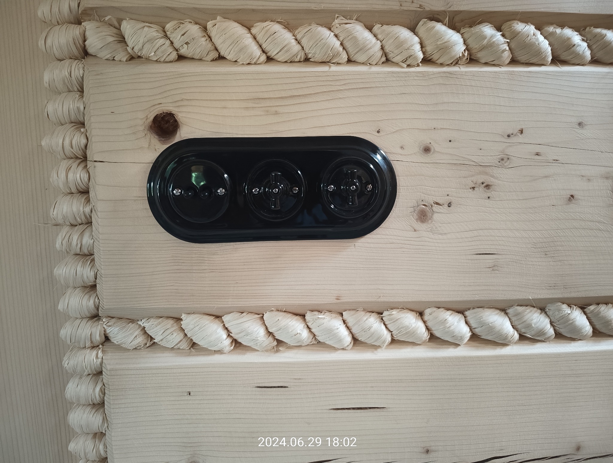 Czarny, potrójny włącznik światła zamontowany na jasnej, drewnianej ścianie z ozdobnymi, jasnymi listwami.