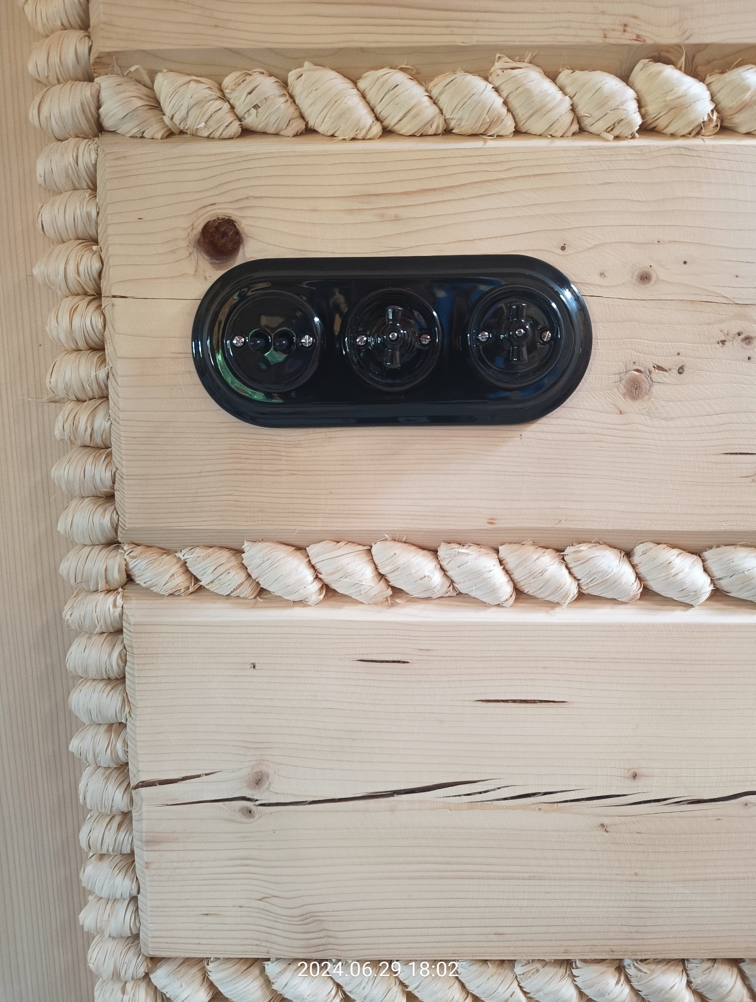 Potrójny, czarny włącznik światła w stylu retro zamontowany na drewnianej ścianie zdobionej dekoracyjnym sznurem z rafii.