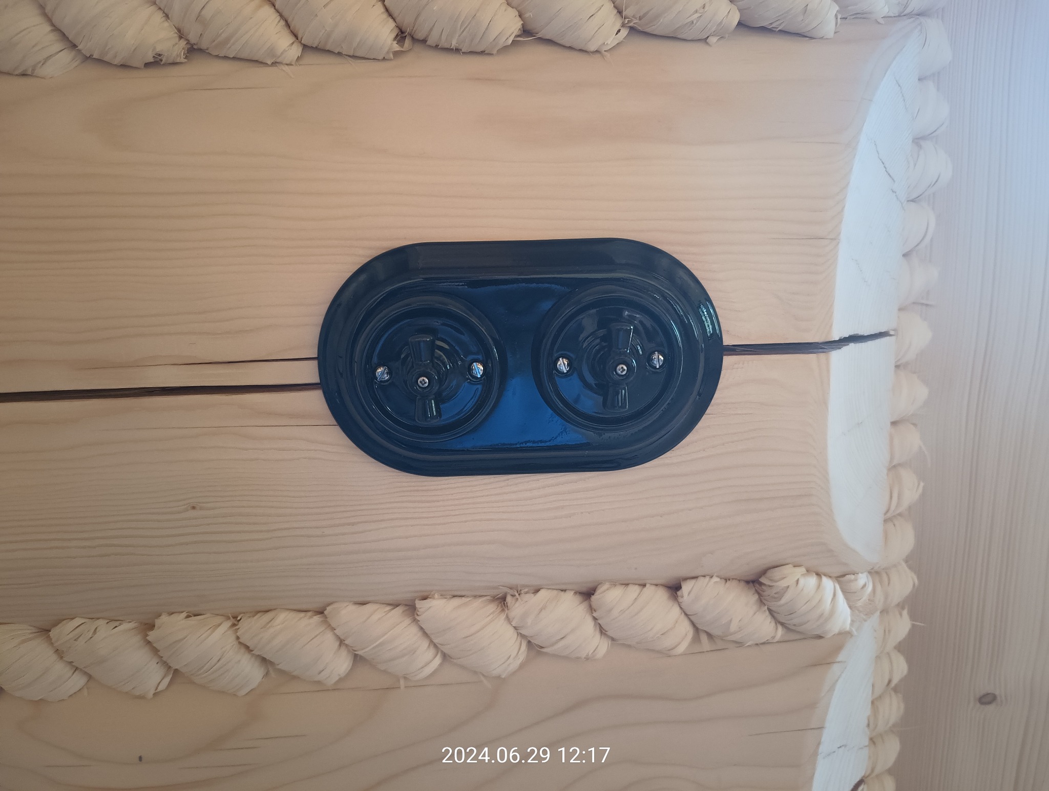 Czarny, podwójny włącznik światła w stylu retro, zamontowany na drewnianej ścianie z ozdobnym wykończeniem w formie jasnej plecionki.