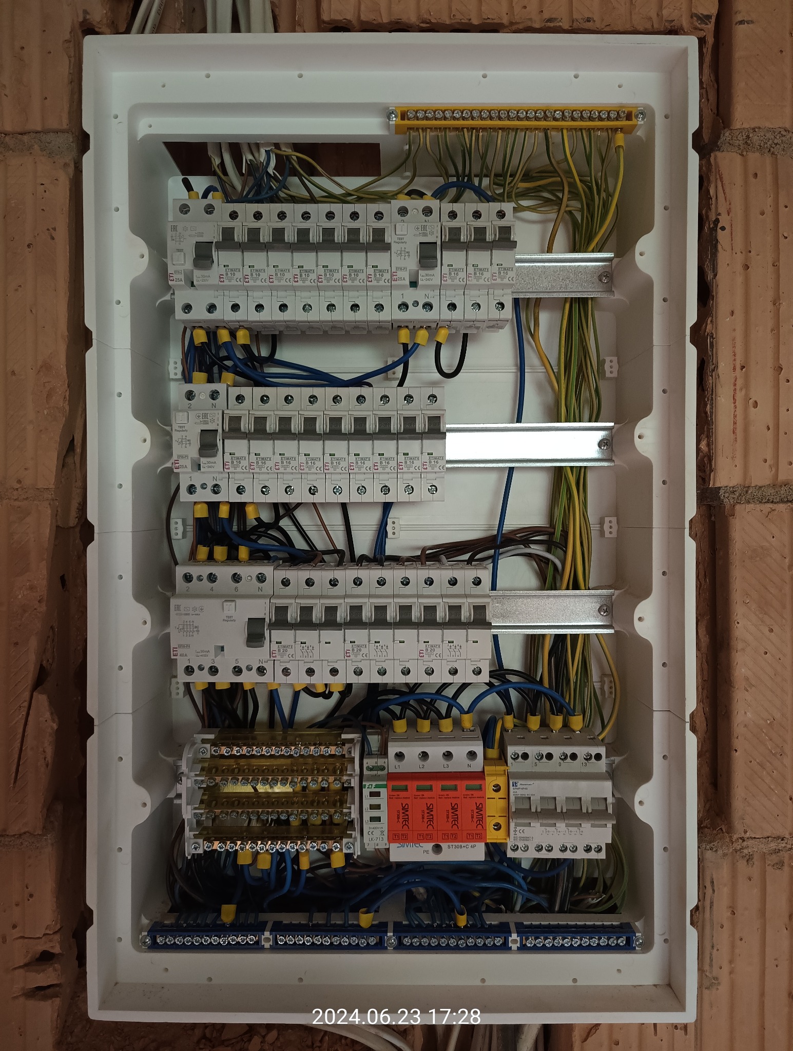 Rozdzielnica elektryczna z zamontowanymi bezpiecznikami, zabezpieczeniami przepięciowymi i okablowaniem, umieszczona w białej obudowie na tle ściany z cegły.