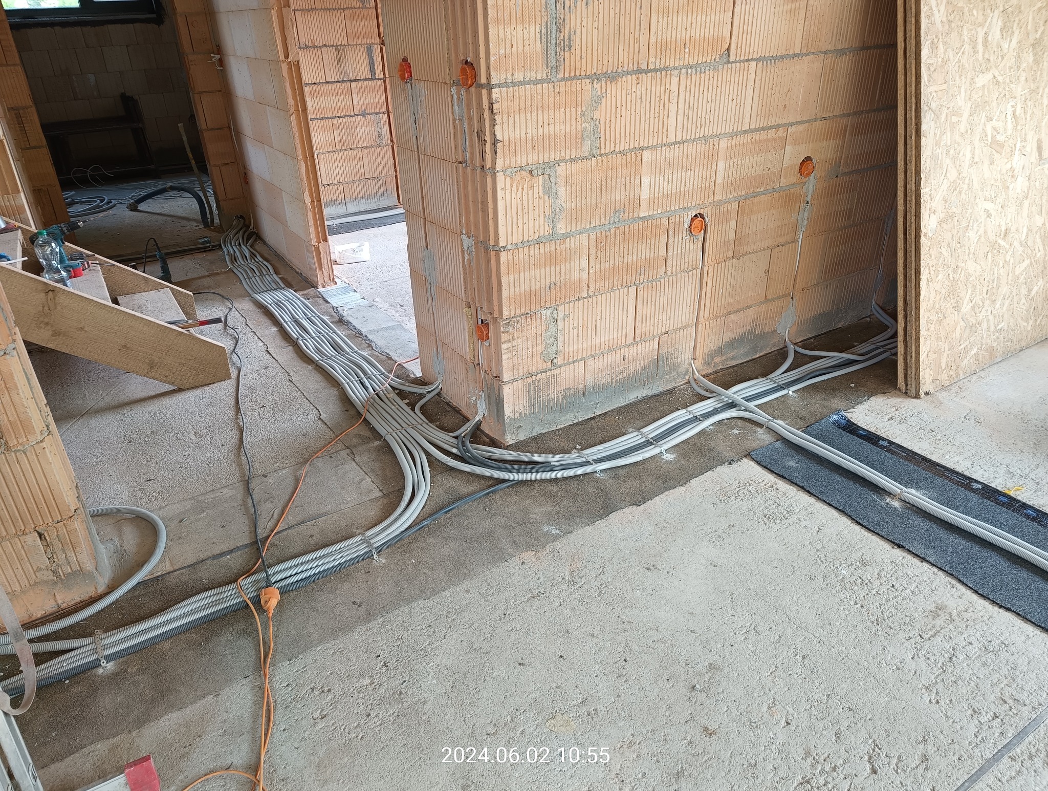 Szereg szarych kabli elektrycznych ułożonych na surowym betonowym podłożu wewnątrz budynku w trakcie budowy, widoczne pomarańczowe puszki instalacyjne w ścianach z cegły dziurawki.