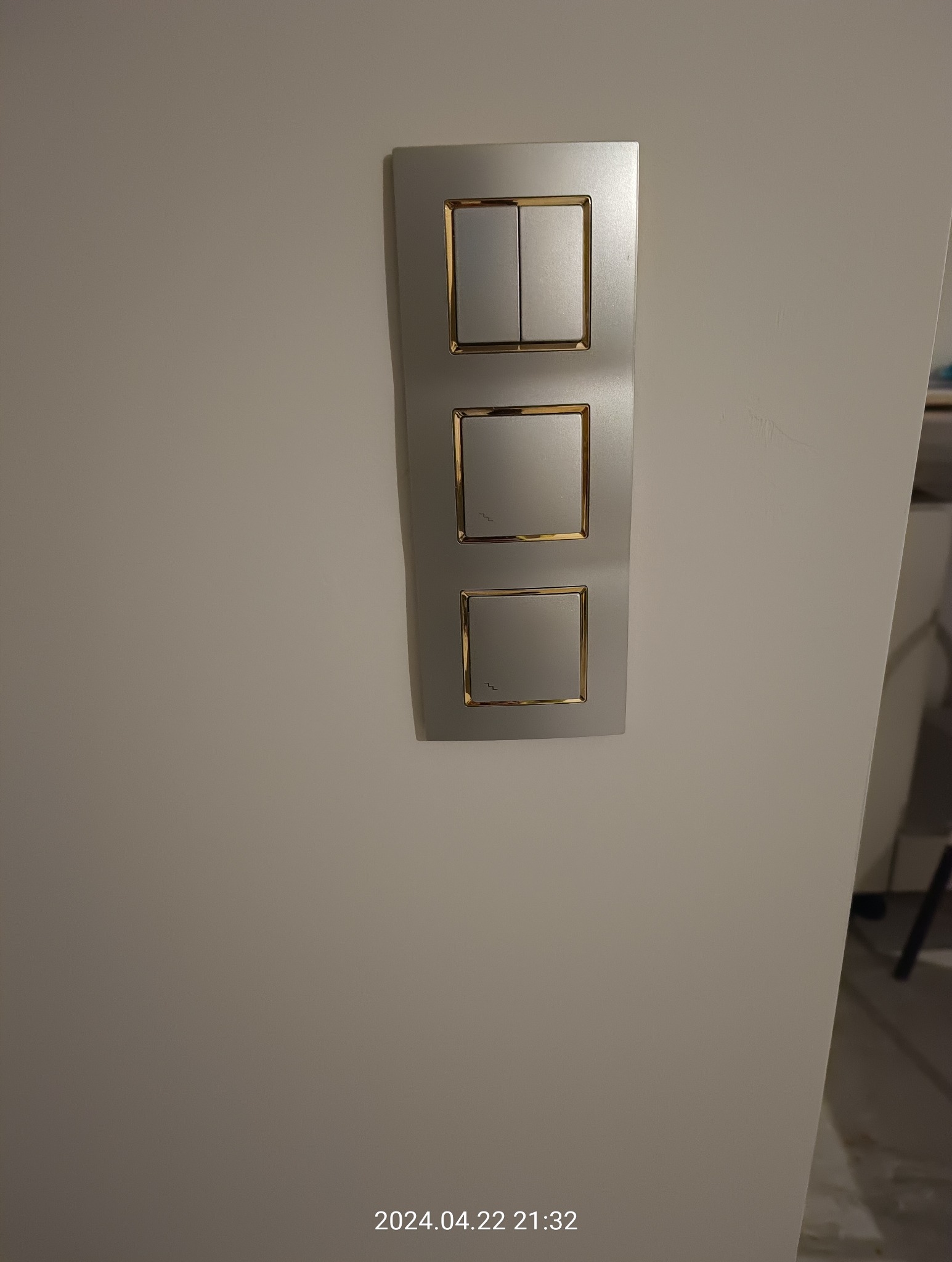 Panel z trzema włącznikami światła w srebrnej ramce z złotymi akcentami, zamontowany na jasnej ścianie. Jeden włącznik dwuklawiszowy, dwa pojedyncze.