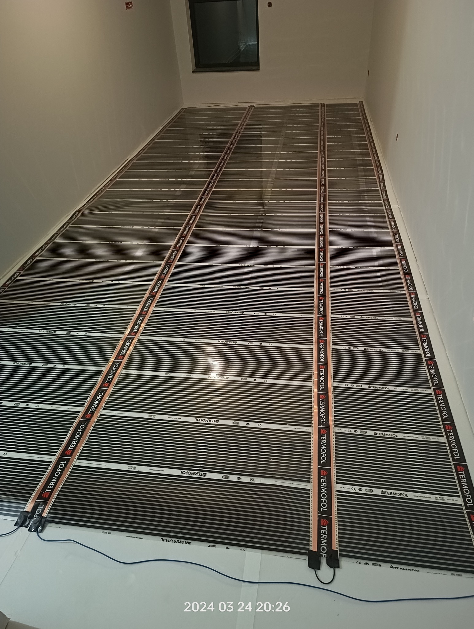 Rozłożona mata grzewcza Termofol na podłodze w trakcie montażu, widoczne białe ściany i okno w tle.