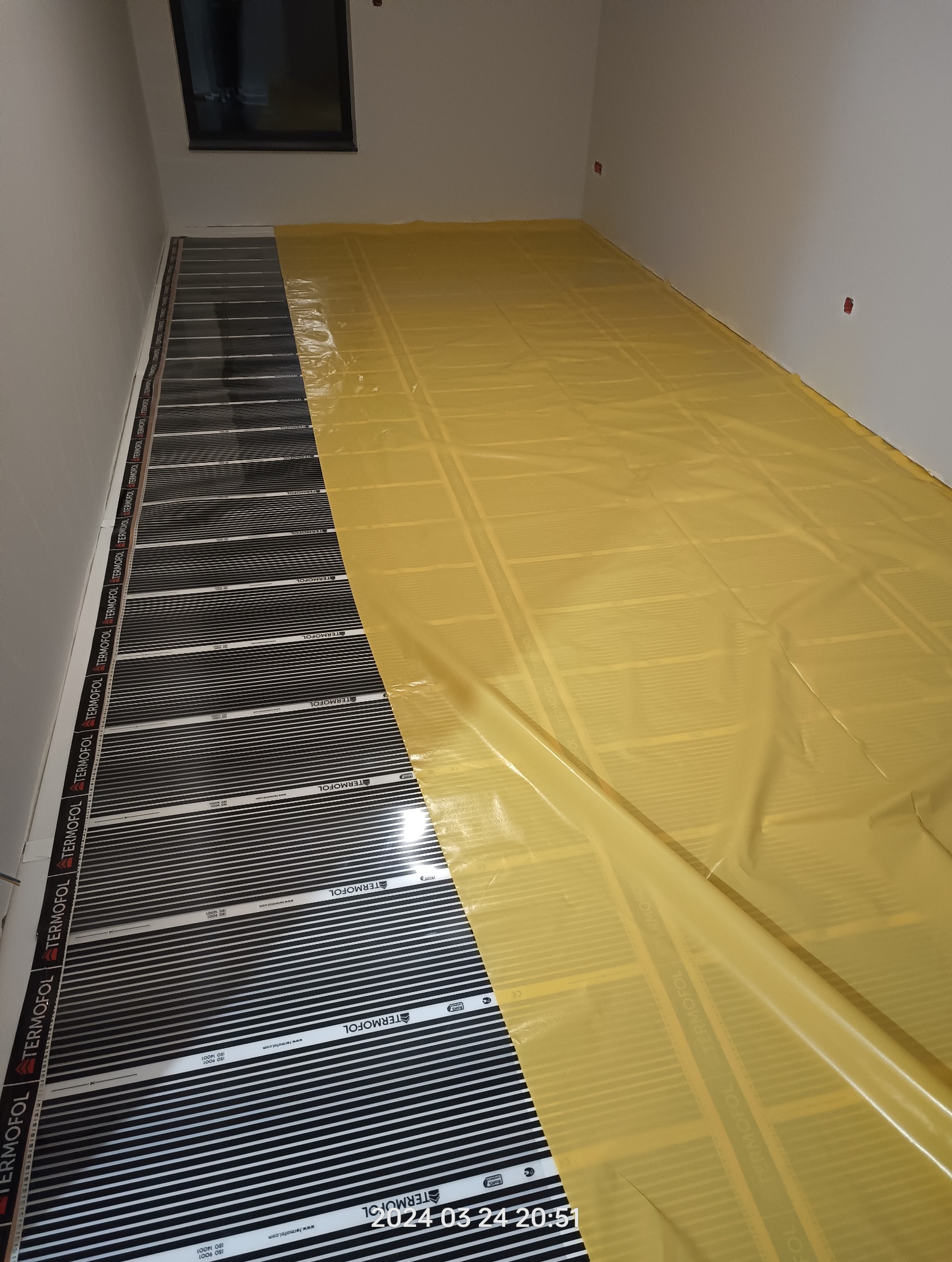 Podłogowe ogrzewanie elektryczne w trakcie montażu, widoczne czarne maty grzewcze Termofol częściowo przykryte żółtą folią ochronną, przygotowane do dalszych prac wykończeniowych.