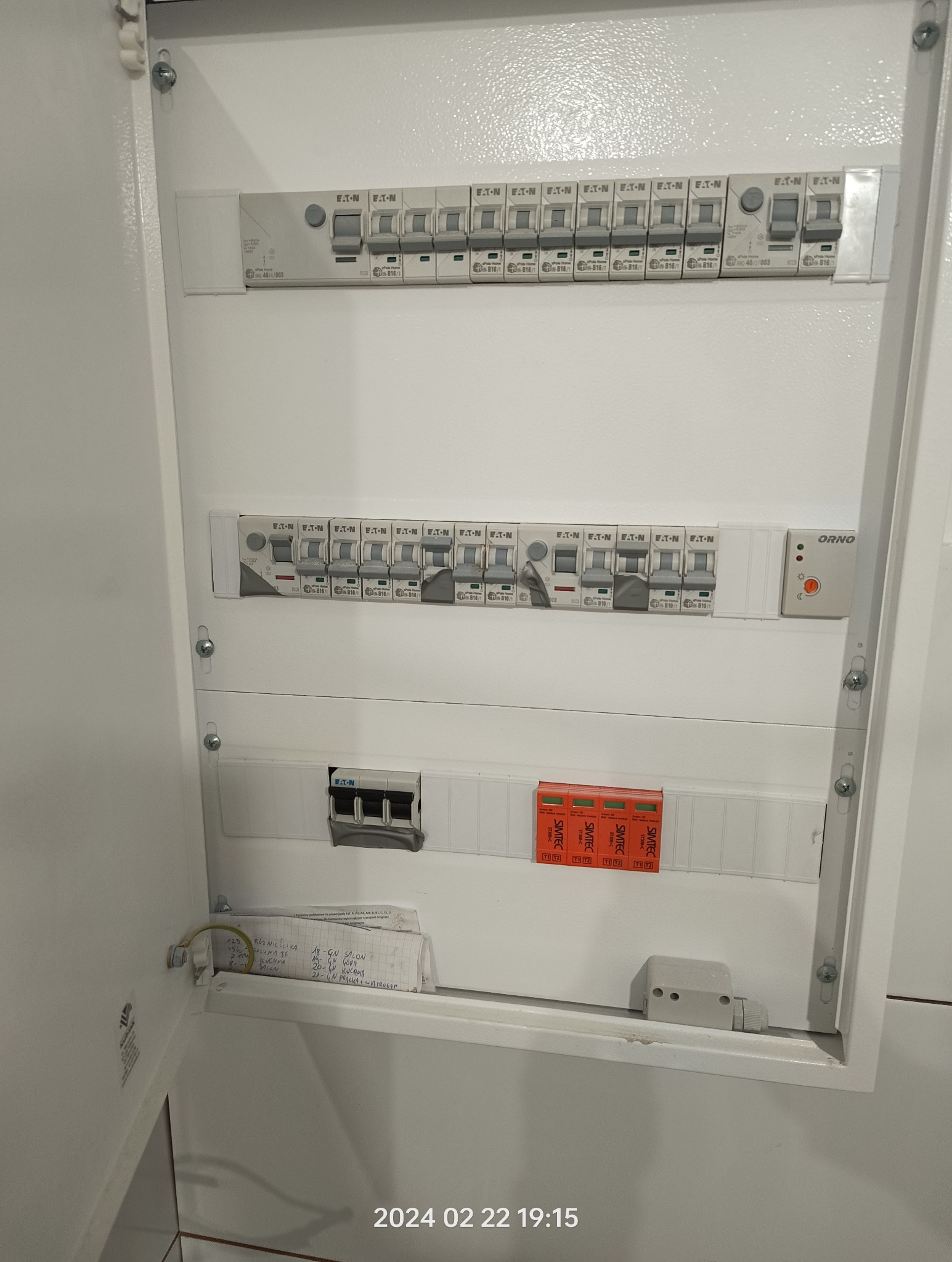 Otwarta skrzynka elektryczna z bezpiecznikami EATON i ORNO, widoczne zabezpieczenia SIMTEC, notatki z rozpisanymi obwodami na dnie skrzynki, białe wnętrze.