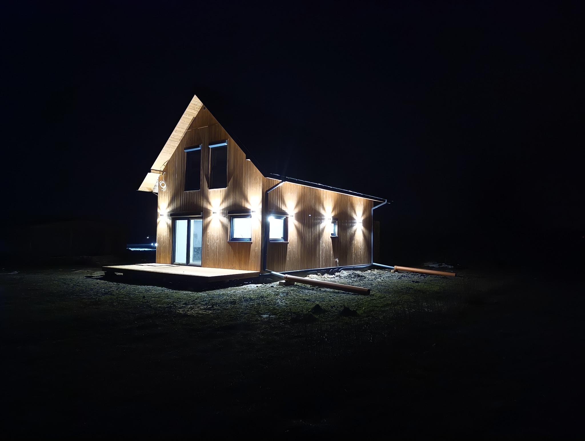 Nowoczesny dom z drewnianą elewacją oświetlony zewnętrznymi lampami w nocy, widoczne rury odprowadzające wodę.