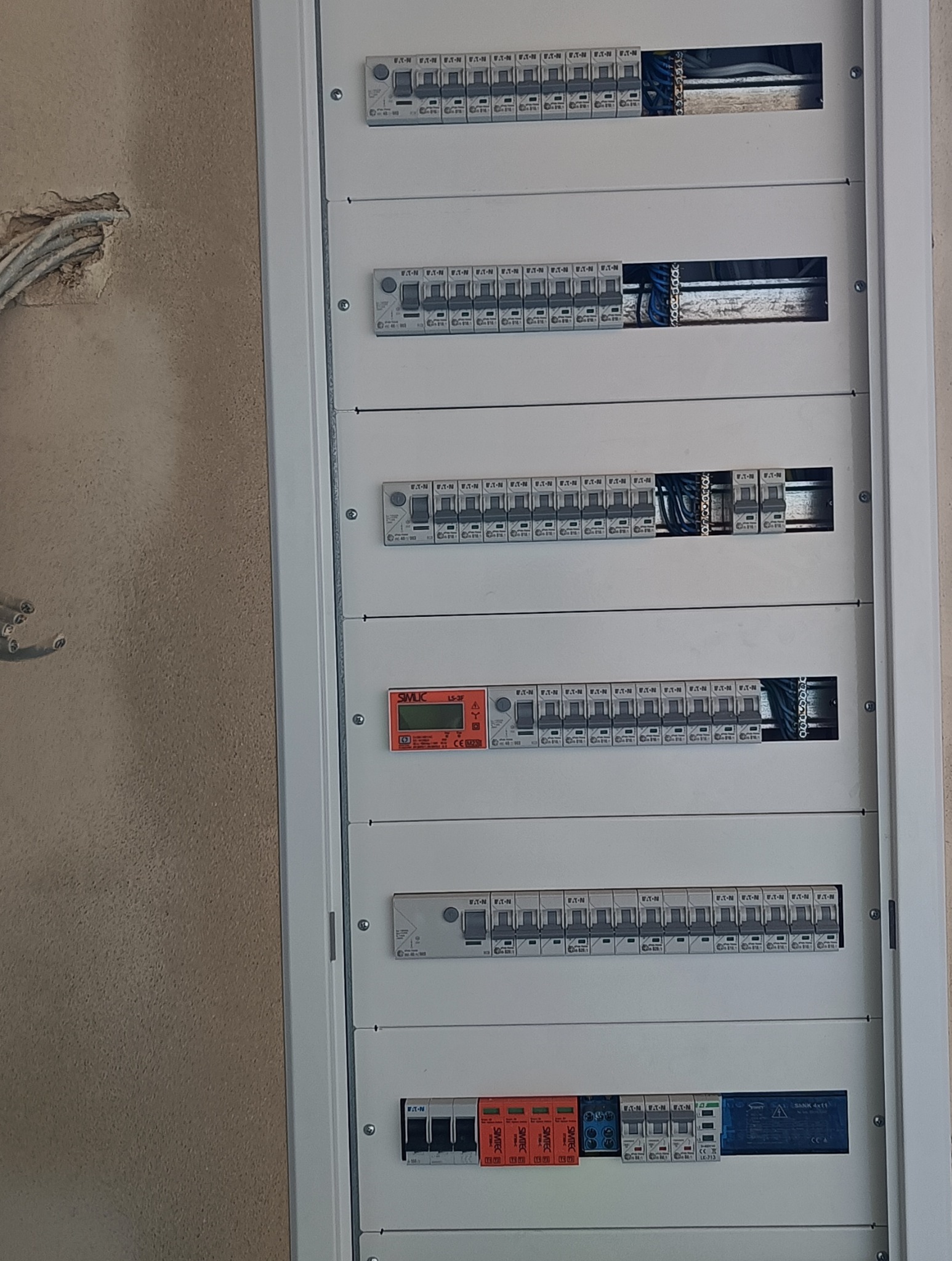 Rozdzielnica elektryczna z widocznymi bezpiecznikami i przewodami, zamontowana na ścianie z surowym wykończeniem. Widoczny licznik Simlic LS-3F. Część bezpieczników jest opisana marką Eaton.