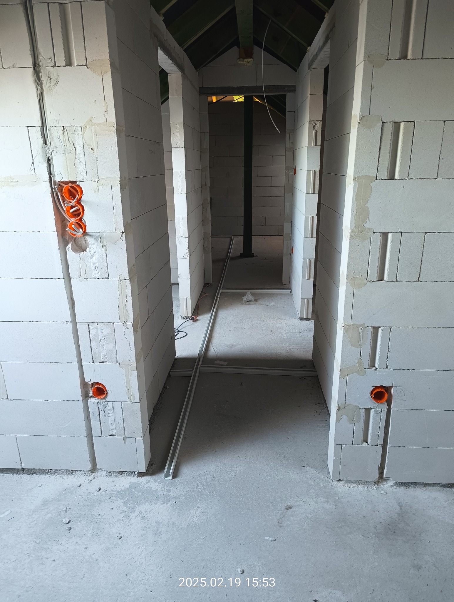 Wnętrze w trakcie budowy z widocznymi pustakami, pomarańczowymi puszkami instalacyjnymi i metalowymi szynami podłogowymi ułożonymi w poprzek korytarza.