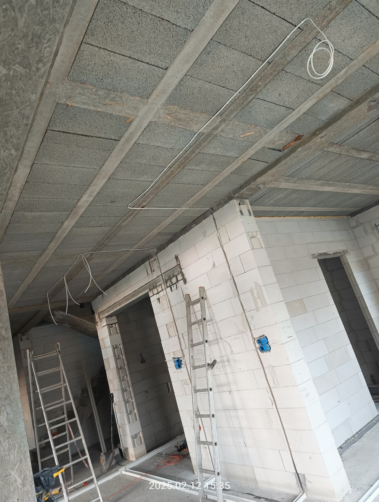 Wnętrze w trakcie budowy z widocznymi ścianami z białych bloczków, drewnianym stropem z widocznymi przewodami elektrycznymi, dwoma aluminiowymi drabinami i niebieskimi puszkami elektrycznymi...