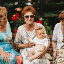 Trzy kobiety siedzą na ławce w ogrodzie, jedna trzyma na kolanach niemowlę z różową opaską na głowie, w tle rozkwitają czerwone róże.