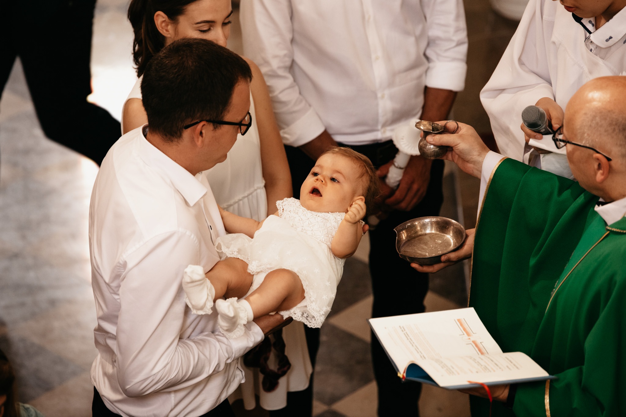 Uroczystość chrztu: niemowlę w białej sukience trzymane przez ojca, ksiądz w zielonej szacie z mikrofonem i naczyniem na wodę święconą.