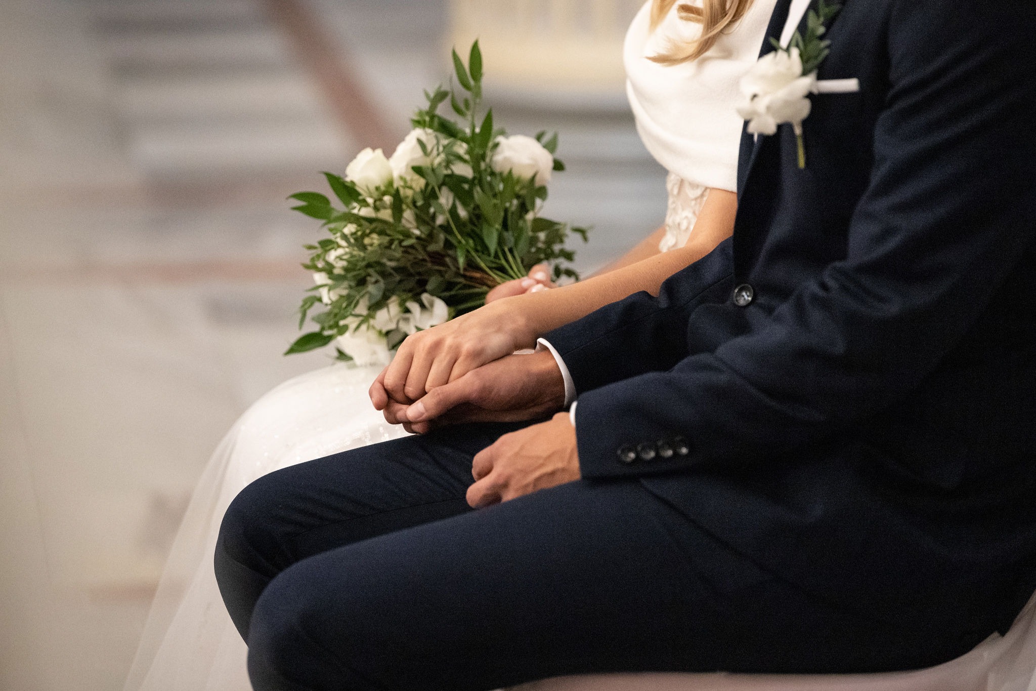 Dłonie pary młodej splecione na kolanach, bukiet białych róż, granatowy garnitur z butonierką, jasna suknia ślubna.