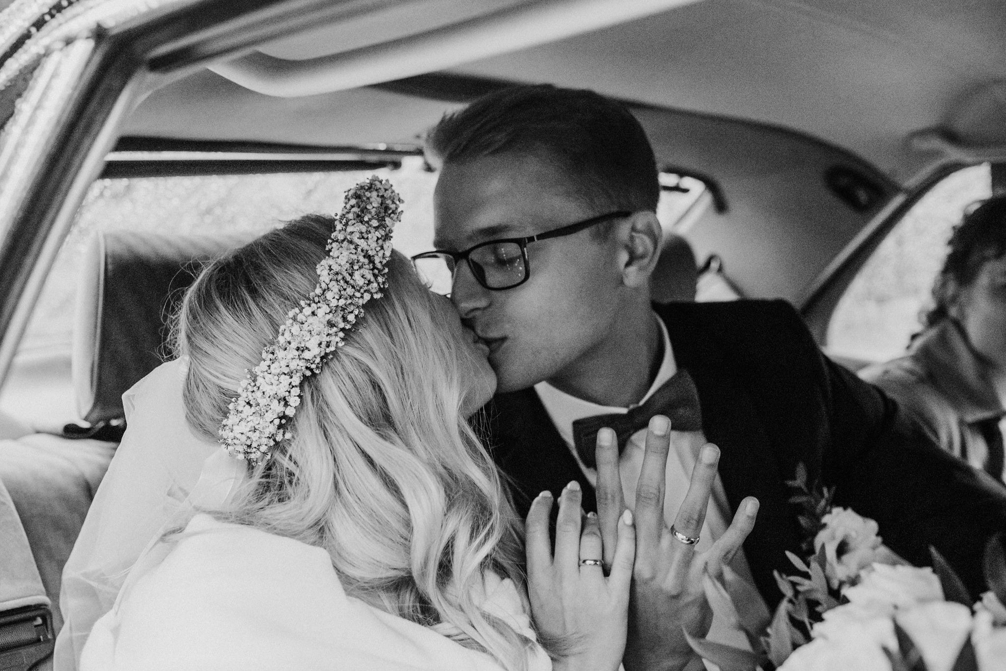 Czarno-białe zdjęcie całującej się pary młodej w samochodzie, panna młoda ma wianek z gipsówki, oboje pokazują obrączki, bukiet kwiatów w tle.