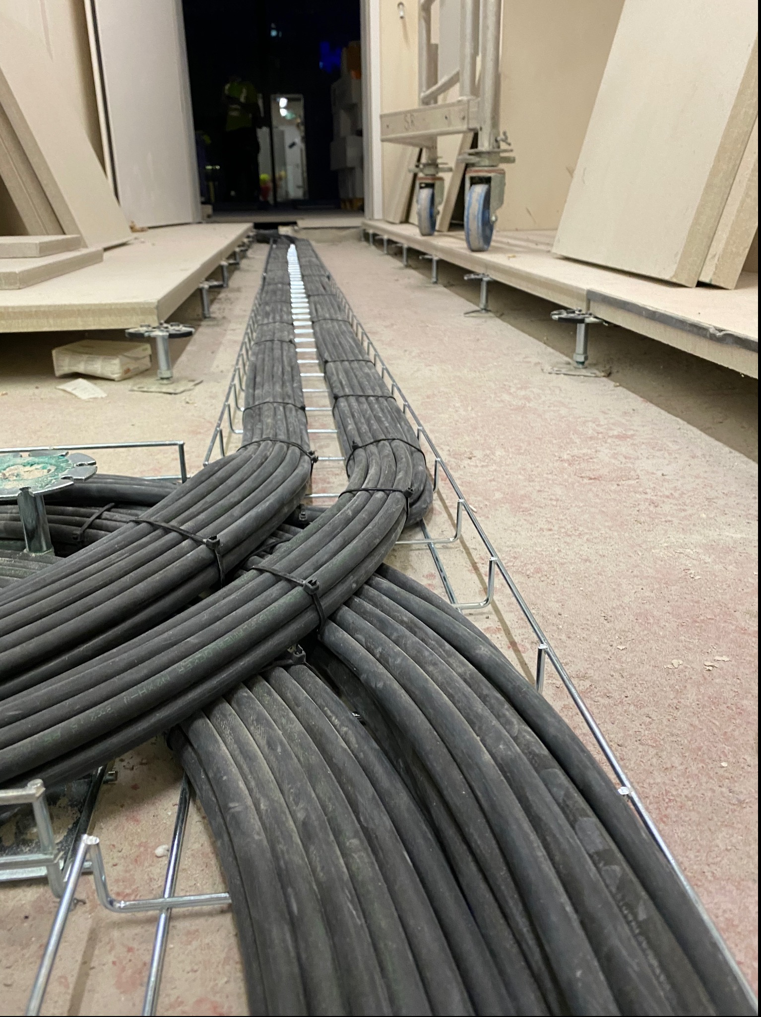 Rząd ułożonych kabli elektrycznych w czarnych osłonach, spiętych opaskami, prowadzonych w metalowej korycie pod podłogą techniczną w nowo budowanym obiekcie.