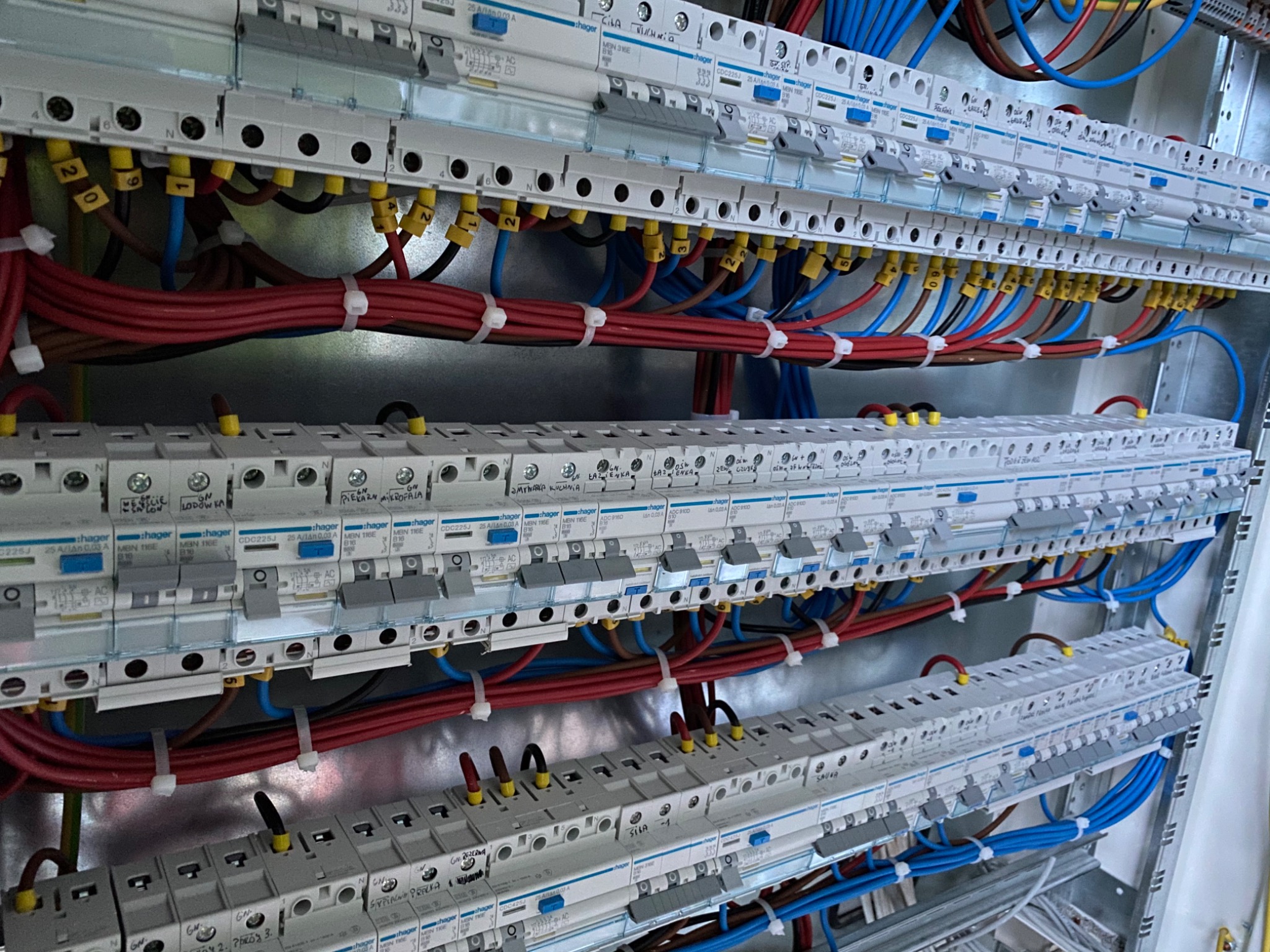 Rozdzielnica elektryczna z zainstalowanymi bezpiecznikami Hager, widoczne przewody w kolorach czerwonym, niebieskim i brązowym, uporządkowane kable spięte białymi opaskami zaciskowymi.