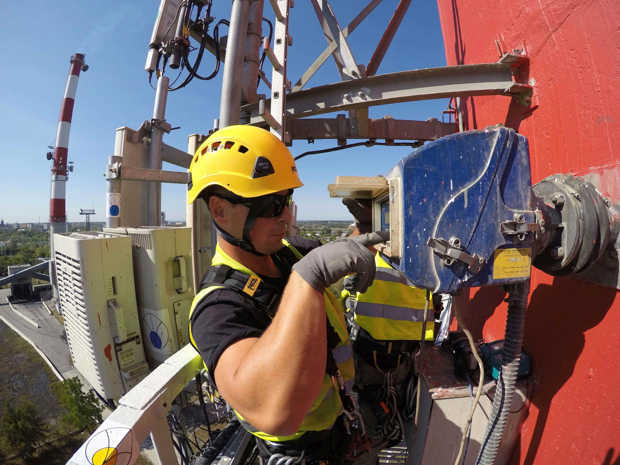 Pracownik w kasku Petzl, na wysokości, używa specjalistycznego urządzenia do obróbki czerwonej, cylindrycznej konstrukcji, prawdopodobnie komina, z widocznym miastem w tle.